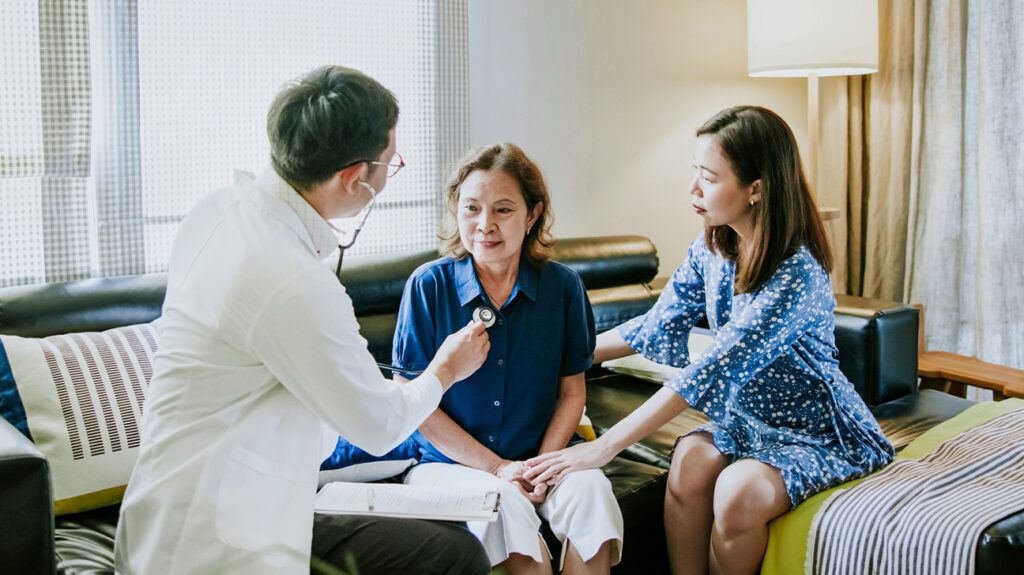 Doctor listening to a woman's heart with a stethoscope. Another woman is sat next to her and supporting her.