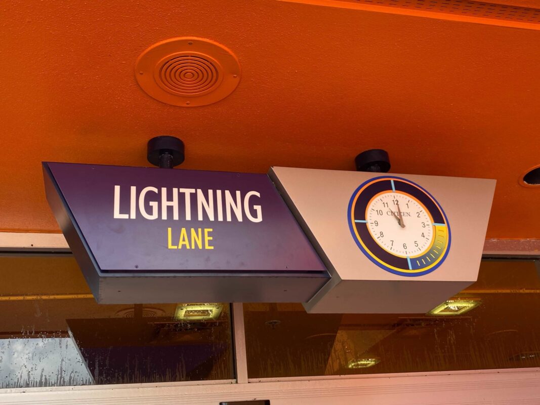 A sign reading "Lightning Lane" next to a circular clock displaying the current time, providing a perfect spot for your daily recap. The sign is mounted under an orange ceiling.