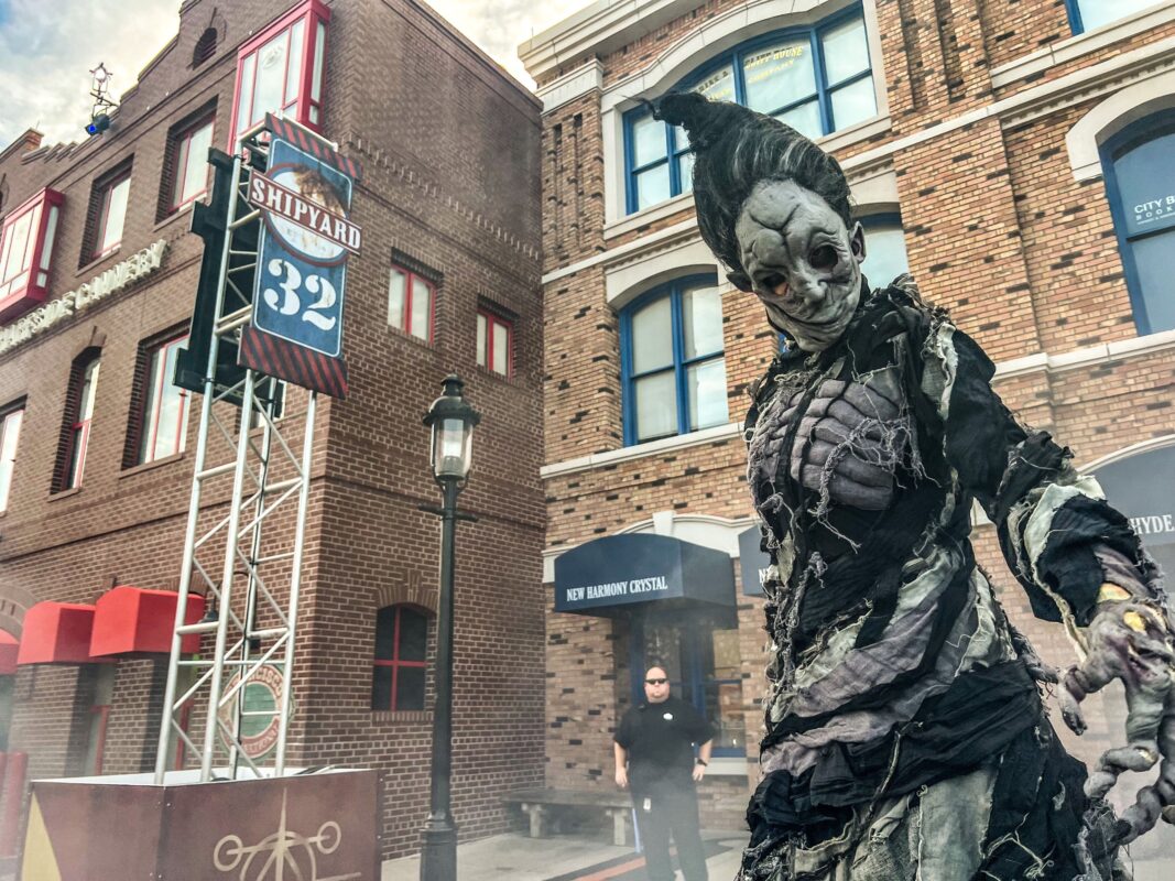 A person in an elaborate, eerie costume stands in front of a brick building with signs reading "Shipyard 32" and "New Harmony Crystal." Another individual lingers near the entrance, both shrouded in an air of mystery.