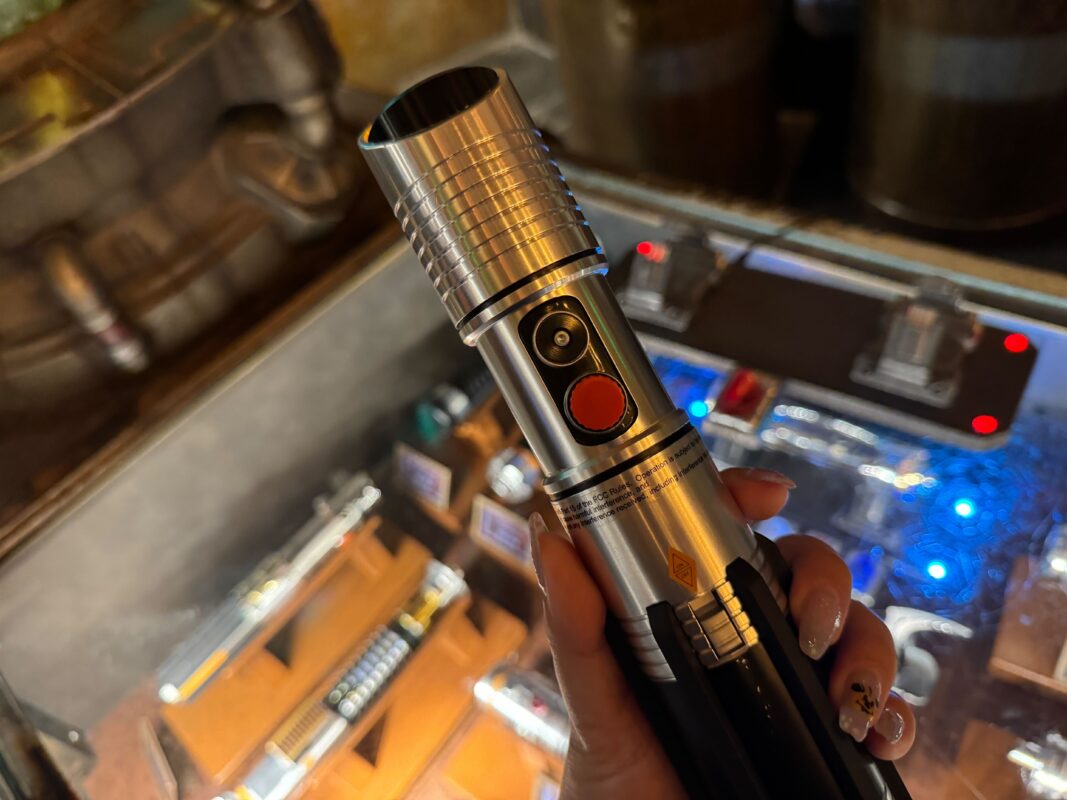 A person holds a silver and black lightsaber hilt with buttons, displayed over a case with various parts, reminiscent of the technology aboard a galactic starcruiser.