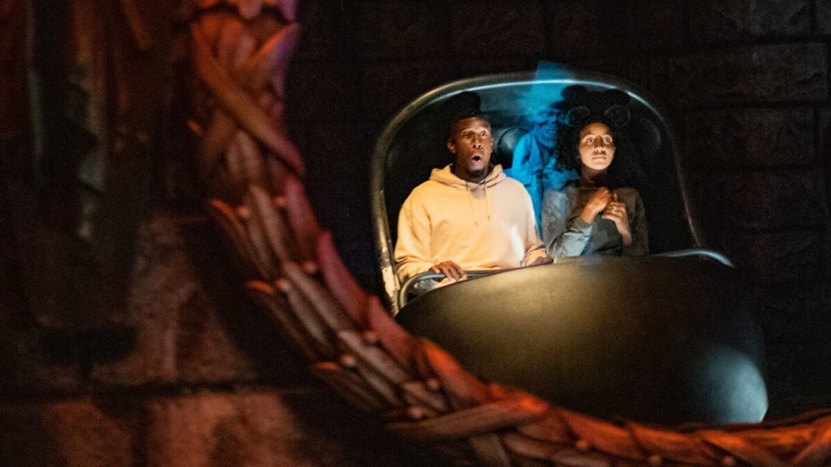 Two individuals are in a dark amusement park ride, sitting closely together and displaying expressions of surprise. The scene is dimly lit with a woven, circular frame partially visible in the foreground, suggesting the presence of hitchhiking ghosts adding to the eerie atmosphere.