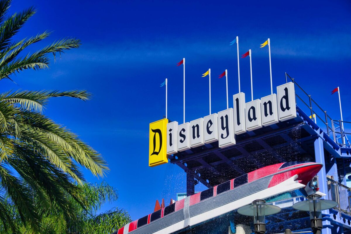 Sign reading "Disneyland" with small flags on top, against a bright blue sky. Palm tree leaves and part of a red and white monorail are seen in the foreground, reminding guests of the Summer Ticket Discounts available.