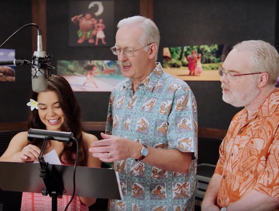 Three individuals stand in a recording studio, with one woman reading a script and two men next to her. A microphone is in front of them, and there are animated images on the wall behind them.