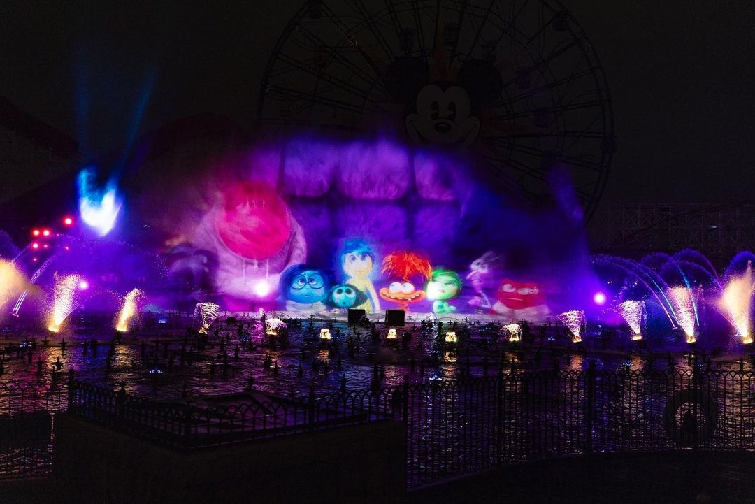 Nighttime water show with colorful projection of animated characters on mist, vibrant fountains, and a large ferris wheel in the background, partially hidden by the display. Tune in for the daily recap to relive every mesmerizing moment.