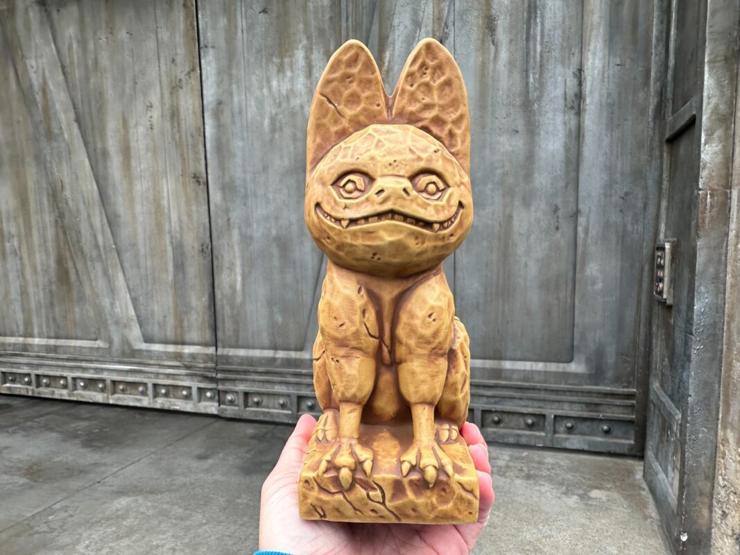 A wooden sculpture of a whimsical loth-cat with a smiling face and large ears, held in a hand against a grey wooden door background.