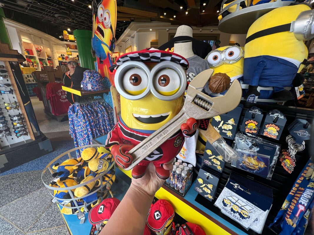 A stuffed Minion toy dressed in red overalls with goggles, holding a large toy wrench, is displayed in a store. Surrounding it on the shelves are various Minion merchandise items, including an adorable Minion popcorn bucket.