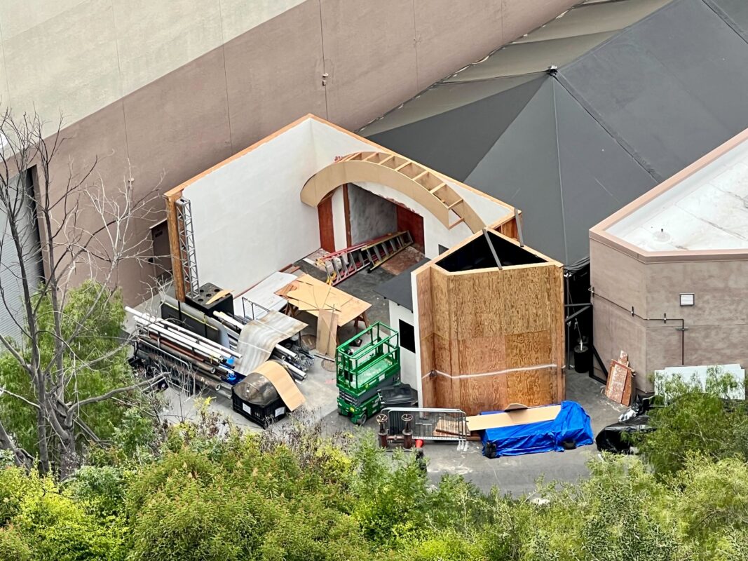 Aerial view of a construction area with a partially built structure. Various building materials and equipment, including scaffolding and a green forklift, are scattered around the site.
