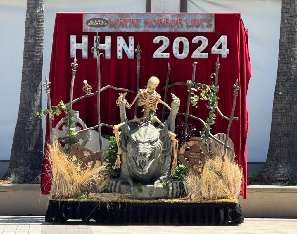 Decorative display for "HHN 2024 - where horror lives," featuring a skeleton, gargoyle, gravestones, and dried plants against a red draped backdrop.
