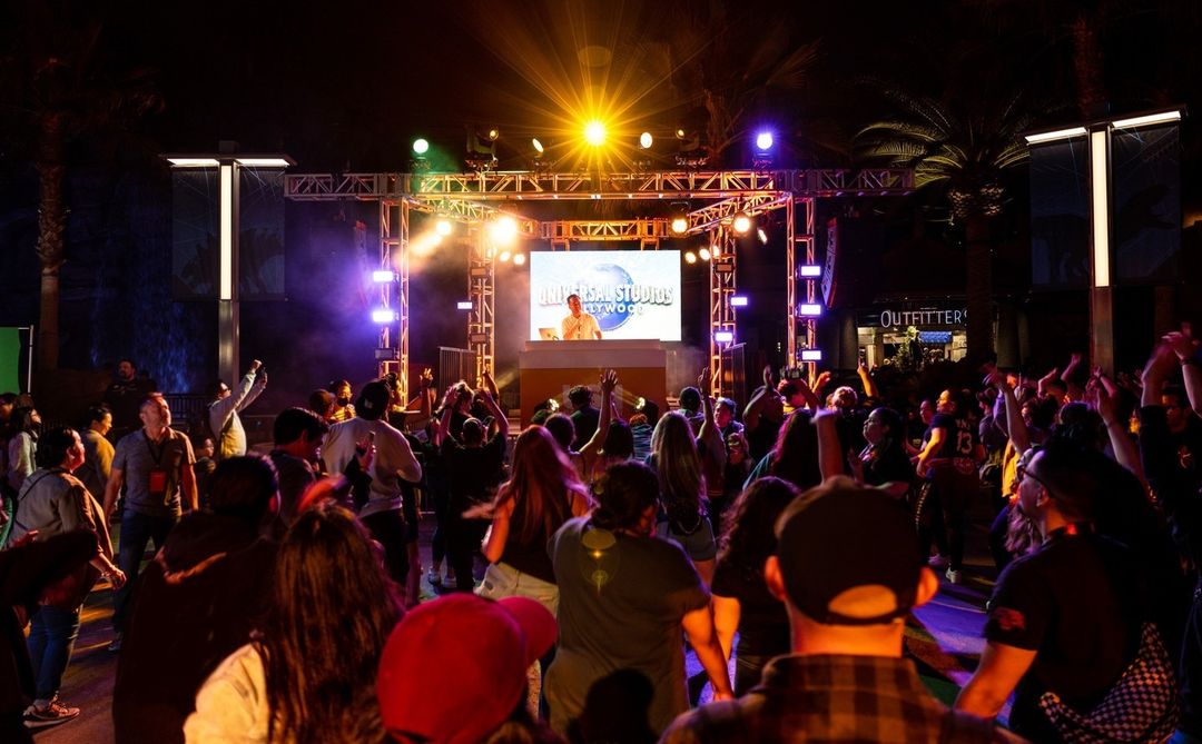 A crowd of people dance and raise their hands at an outdoor concert with colorful stage lights and a large screen displaying a performer.