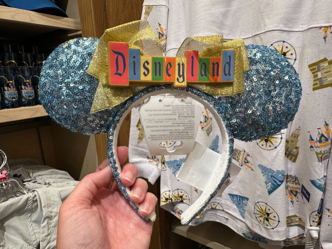 A blue sequined Disneyland Ear Headband with a gold bow and a "Disneyland" sign is displayed in the store as a New Arrival. The background shows a white shirt with Disney castle prints.