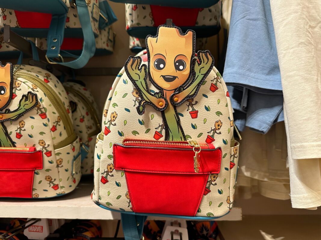 A small backpack featuring a cartoon character with a tree-like appearance, depicted with arms raised. The backpack has a red front pocket and a patterned background with the same character in various poses.