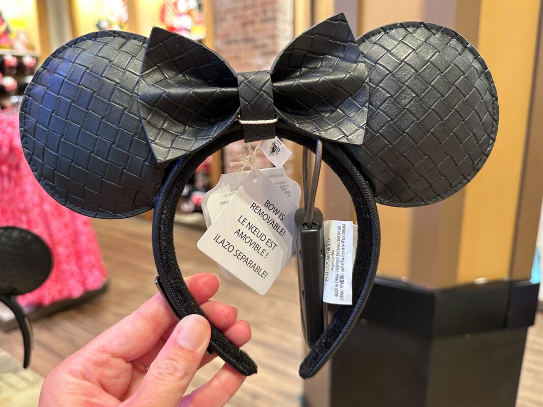 A hand holding a black Loungefly ear headband with a removable Minnie bow in a store. Tags with text are attached to the headband, adding to its charm.
