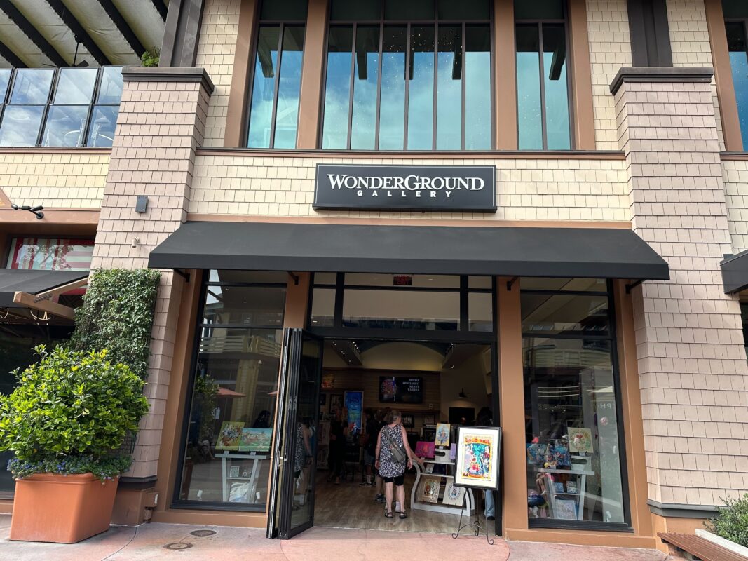 Facade of WonderGround Gallery with large windows and an awning, displaying art pieces. Two women are entering WonderGround Gallery, and there's artwork on easels outside the entrance.