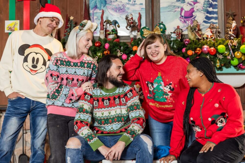 A group of five people wearing festive sweaters and holiday headbands, standing and sitting in a room decorated for Christmas with ornaments and garlands on the wall behind them.