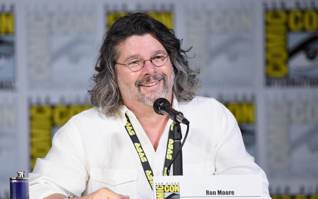 A man with long hair and glasses speaks into a microphone at a panel event. He is smiling and sitting behind a nameplate that reads "Ron Moore." The background features Comic-Con branding alongside the Society of Explorers and Adventurers logo.