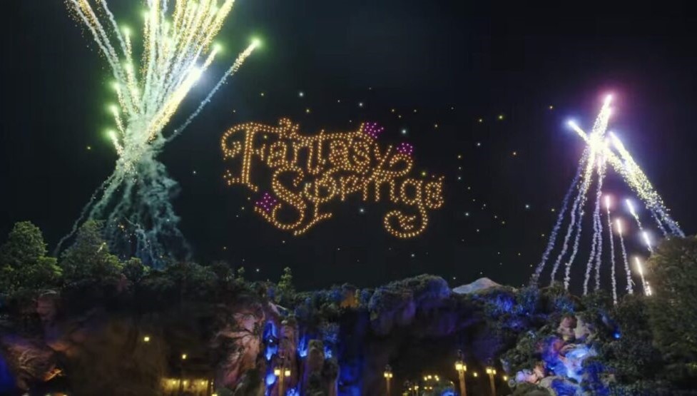 A magical nighttime scene showcases a dazzling blend of fireworks and a drone light display, spelling out "Fantasy Springs" in the sky above an enchanting landscape illuminated with trees and rocks.