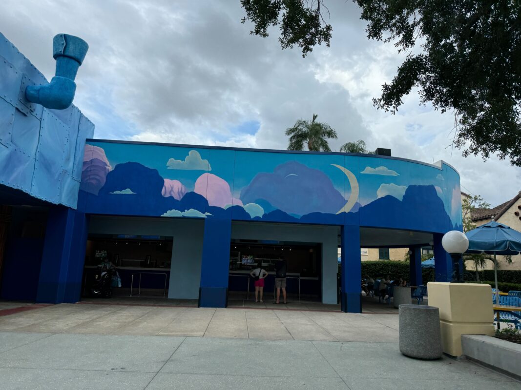 A building with a cloud-themed mural on the upper part, featuring shades of blue and pink, stands under a cloudy sky. Two people are at the entrance of Imagination Cafe, and a tree and lamp post are visible, adding to the dreamy scene.