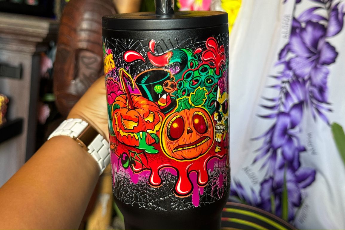 A hand holds a black Lil' Boo Tumbler decorated with colorful, cartoonish Halloween-themed graphics featuring pumpkins, skulls, and splatters.