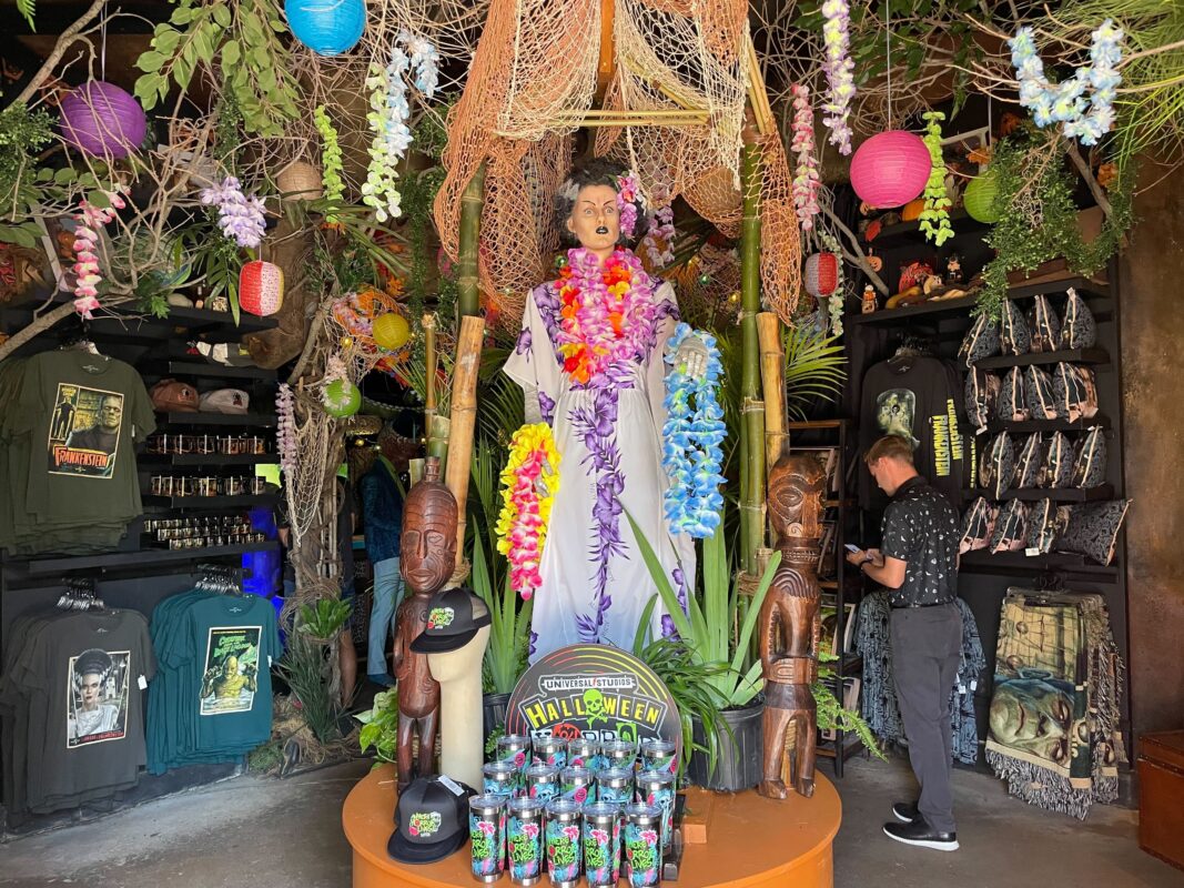 A store display featuring a mannequin adorned with colorful leis, surrounded by tropical decorations, wooden statues, and various merchandise including shirts and bottles. A person stands in the background.