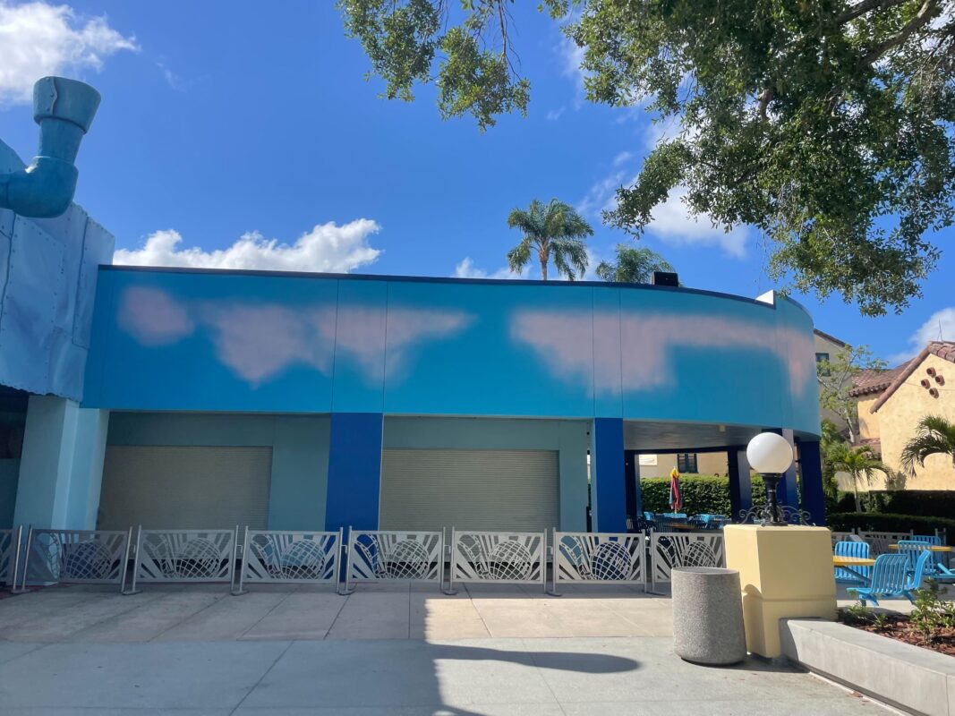 A building with a cloud-themed mural on its wall and closed shutters stands in front of metal barriers and some outdoor seating. The sky above is clear with a few clouds, while trees are visible nearby, hinting at the playful spirit of KidZone Pizza Company.