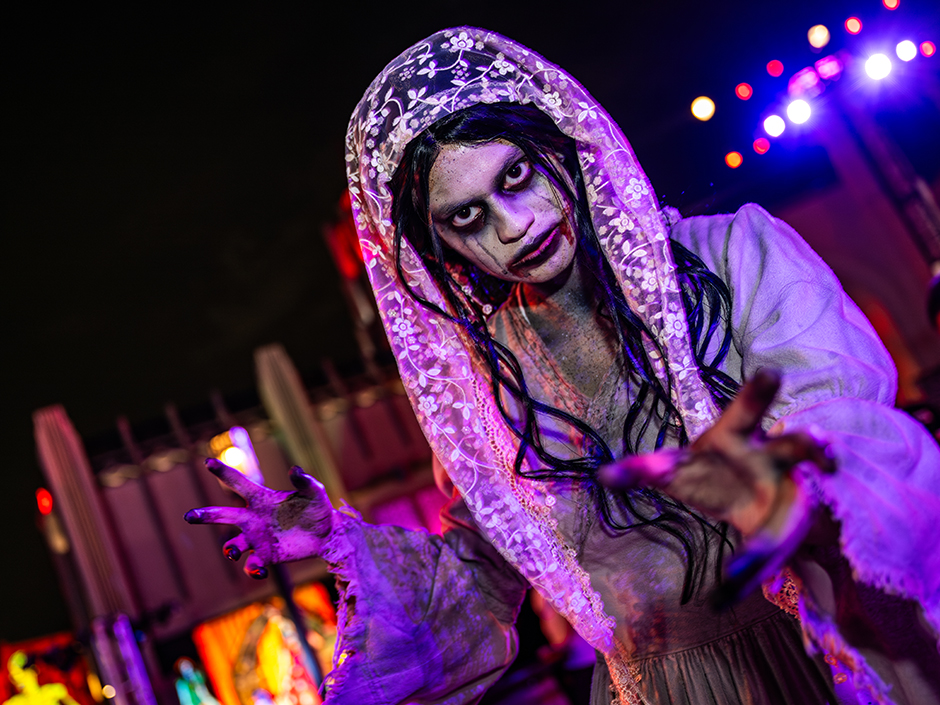 Person in a ghostly costume with white face paint, wearing a veil, long dress, and reaching out with hands. Background features colorful lights and structures.