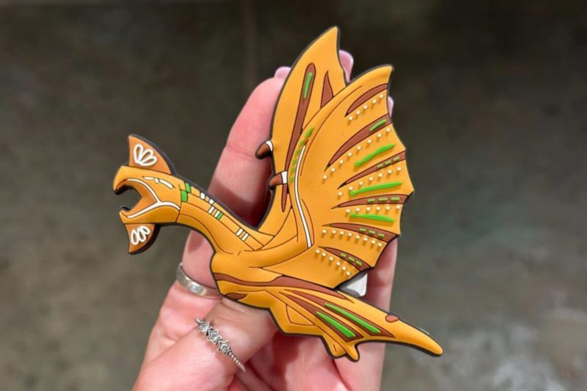 A hand holds a detailed, yellow-orange enamel pin of a dragon with green, white, and red accents. The dragon has outstretched wings and an open mouth, echoing the fierce elegance of a banshee's scream. This mesmerizing pin is sure to become a magnet for compliments.