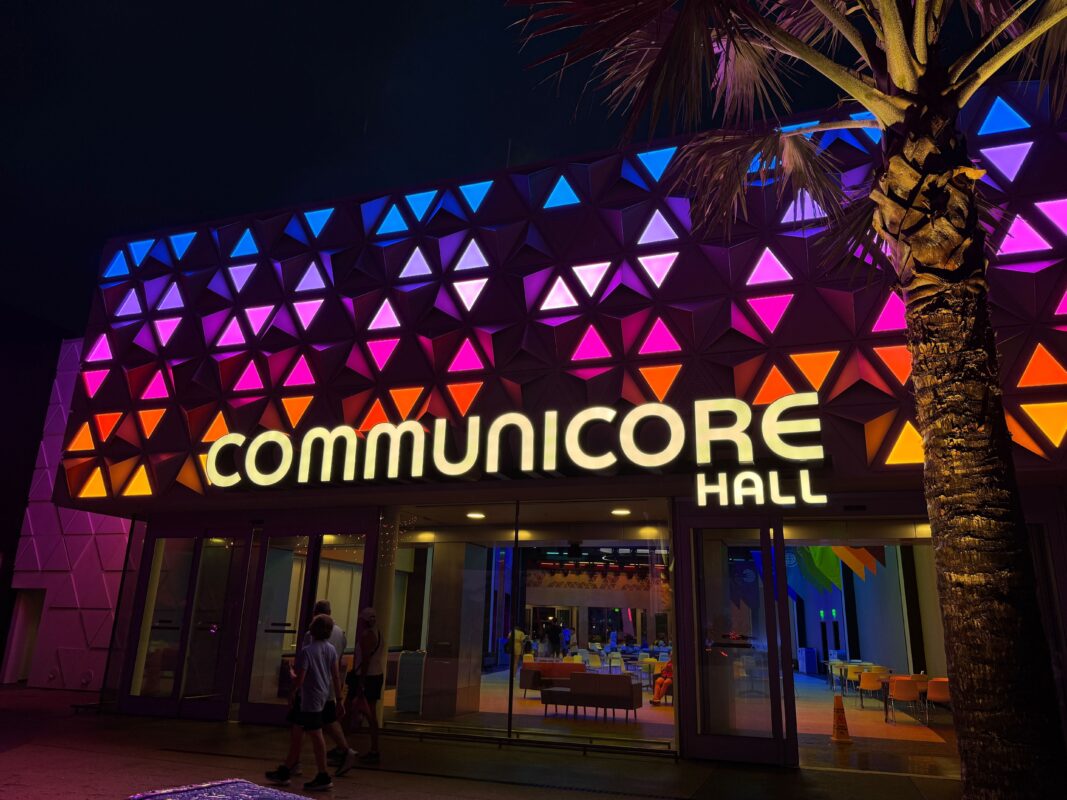 The exterior of CommuniCore Hall at night, illuminated with colorful triangular lights. A tree and a few people are visible near the entrance of CommuniCore Hall.
