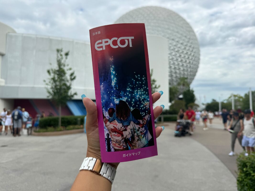 A person holds an Epcot guide map with a blurred view of Spaceship Earth and people walking in the background.