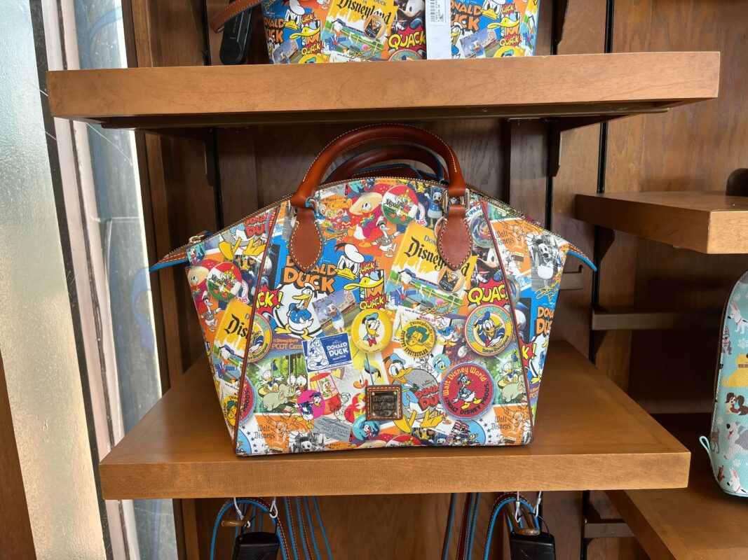 A colorful handbag from the Dooney & Bourke Collection, featuring Disney-themed illustrations including a Donald Duck 90th-Anniversary design, is displayed on a wooden shelf. The bag has brown leather handles and a multicolored collage of various Disney characters and logos, reminiscent of Walt Disney World magic.