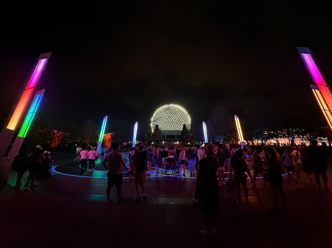 A nighttime scene at an amusement park with illuminated structures and a large spherical building in the background, surrounded by a crowd of people enjoying World Celebration at EPCOT.
