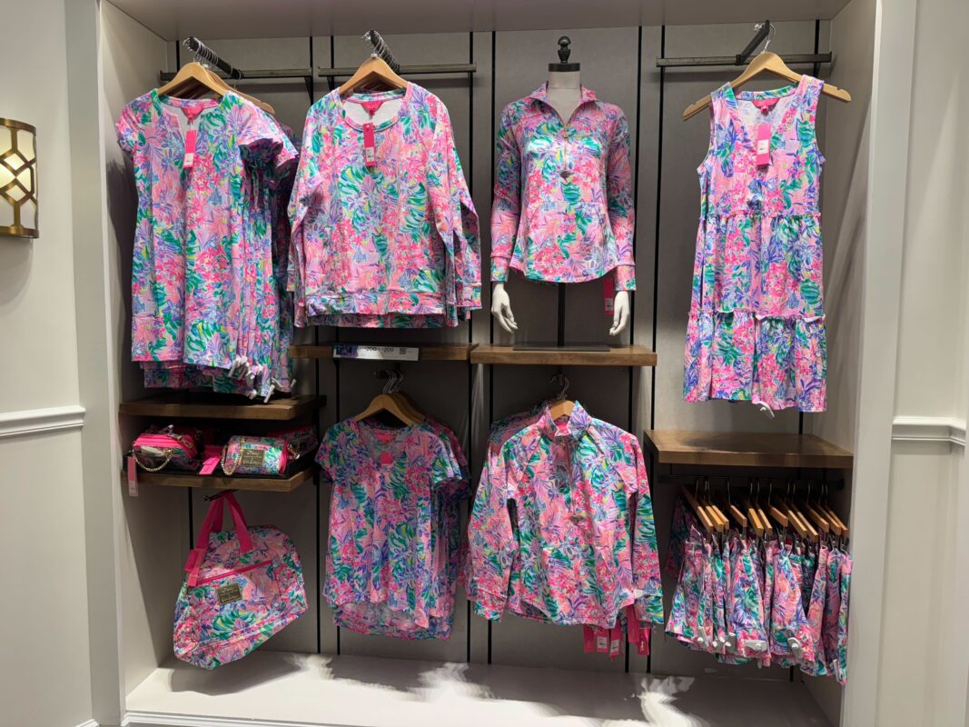 A retail display showcases a collection of pink and green floral-patterned Lilly Pulitzer apparel, including shirts, dresses, and bags, arranged on hangers and shelves.