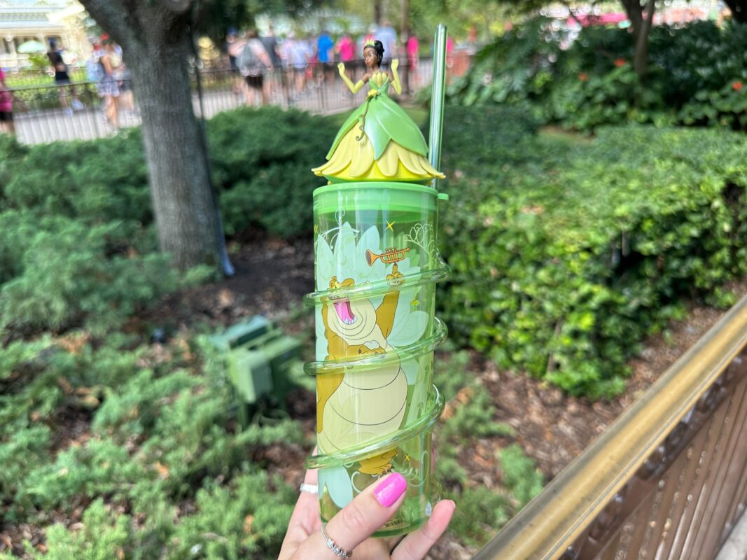 A hand holding a green, princess-themed, spiral cup with a lid featuring a small princess figurine on top and a straw. The background shows greenery and an amusement park setting.