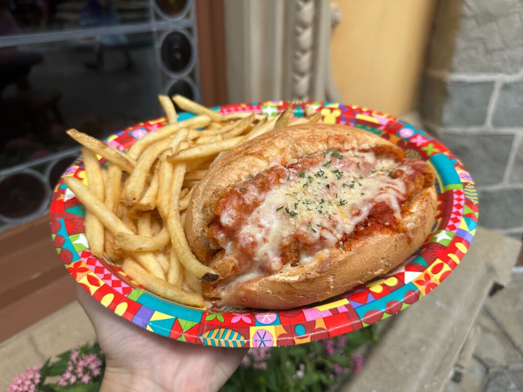 A hand holds a colorful plate with a Chicken Parmesan Sandwich from Pinocchio Village Haus, topped with melted cheese and herbs, alongside a serving of French fries.