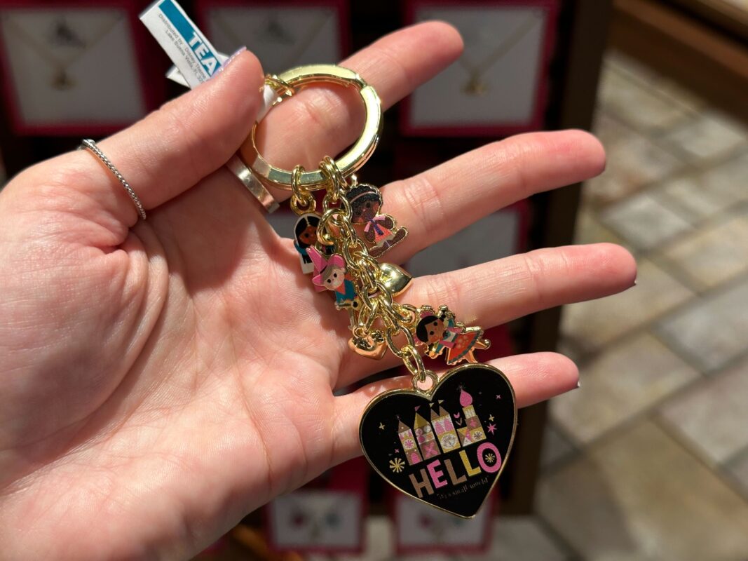 A person holds a keychain featuring a heart-shaped charm with "Hello" text and castle design, along with several smaller character charms.