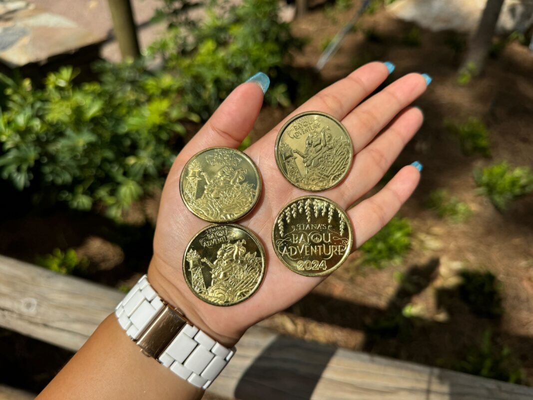 A hand holding four large gold coins, each displaying different intricate designs and text. The person's nails are painted blue, and they are wearing a white wristwatch. Background features greenery.
