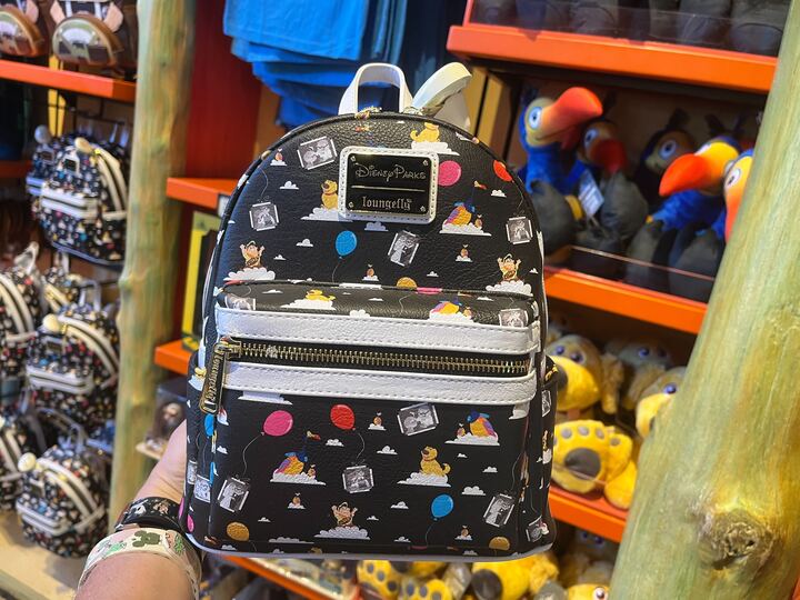 A hand holding a small black Disney-themed backpack with colorful character prints and a white front pocket. Shelves with plush toys are visible in the background.