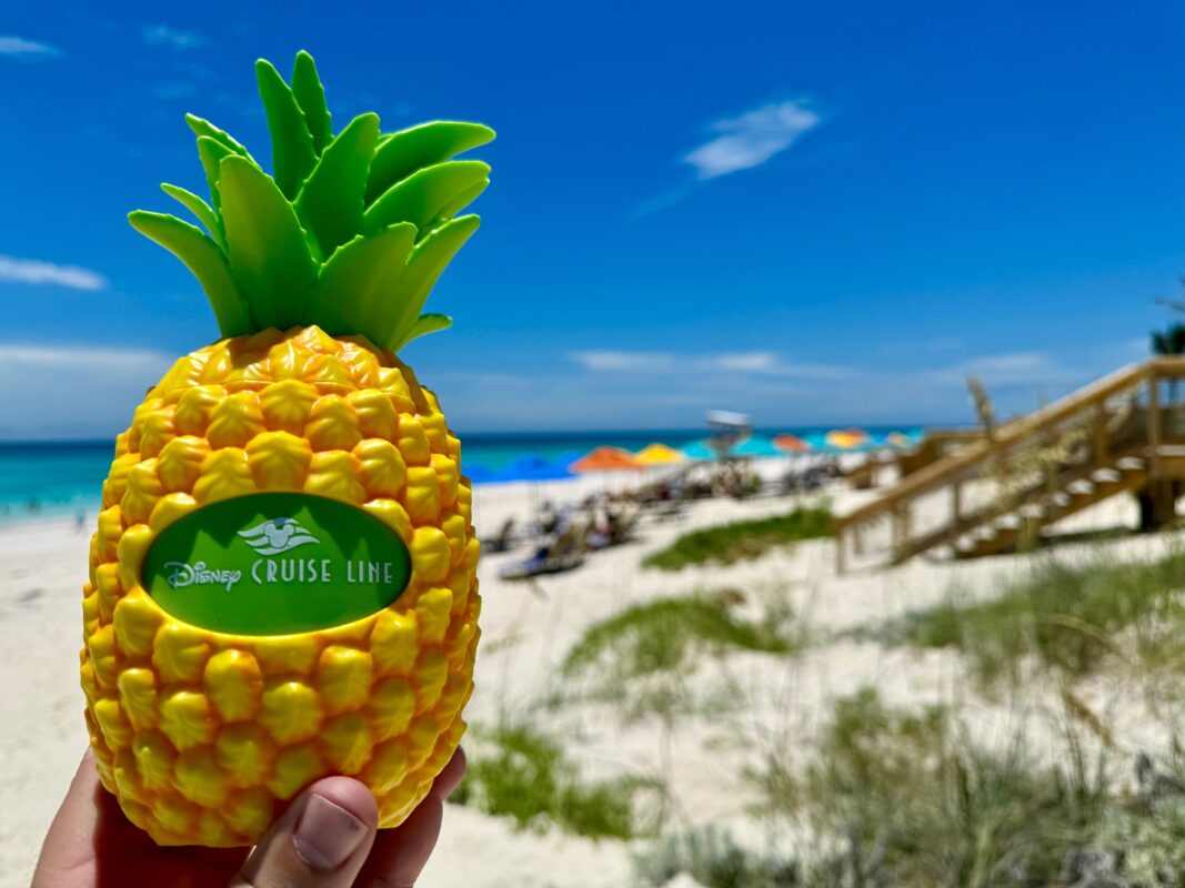 A hand holds a yellow plastic pineapple sipper with a Disney Cruise Line logo. In the background, a sandy beach, colorful umbrellas, and a lifeguard tower are visible under a clear blue sky.