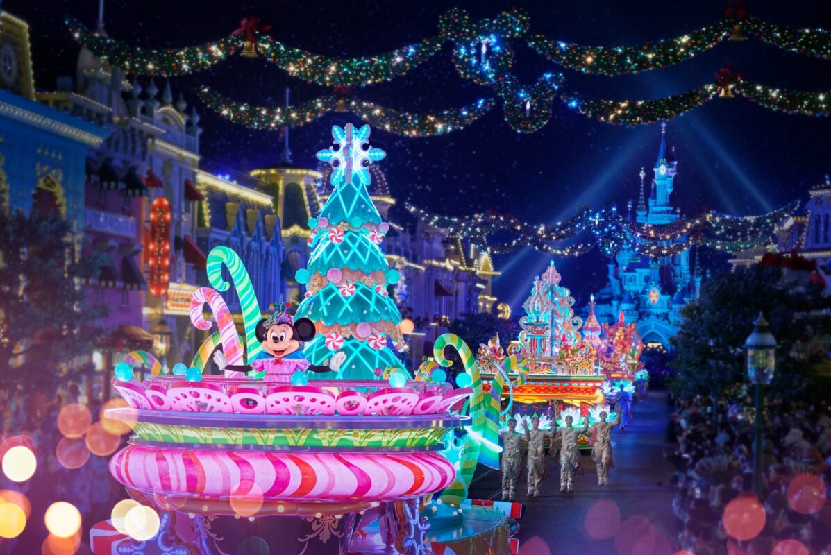 Nighttime parade at a theme park with brightly lit floats, including a Christmas tree themed float featuring a mouse character, surrounded by decorations and garlands.