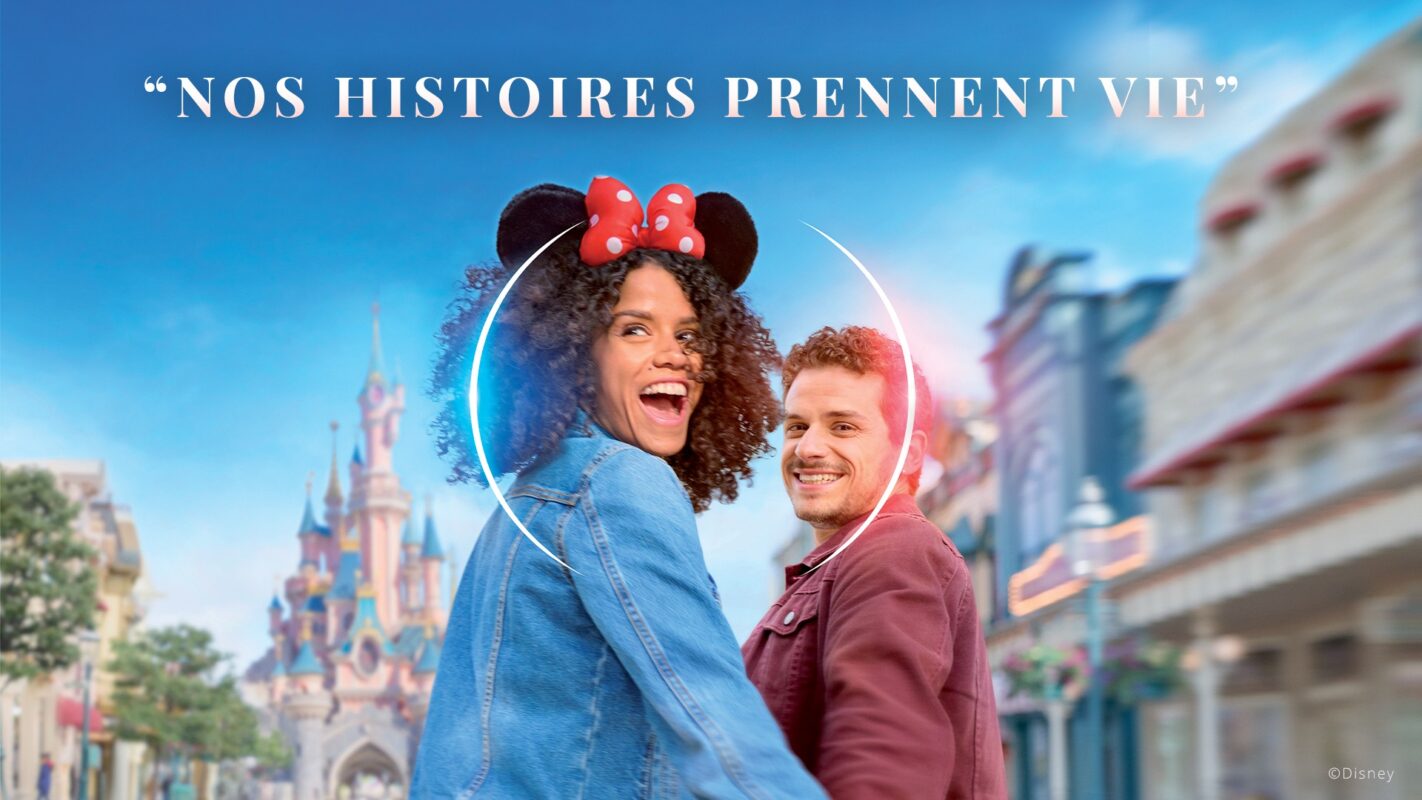 Two smiling individuals, one wearing Mickey Mouse ears, stand in front of a castle at a theme park. Above them are the words "Nos Histoires Prennent Vie.