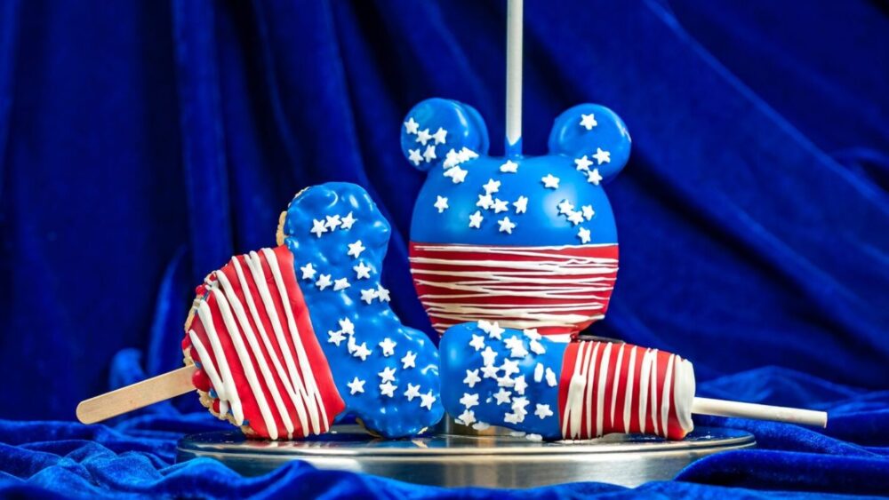 A glossy candy apple and marshmallow pops, all decorated in red, white, and blue with stars, sit on a silver dish against a blue velvet background—perfect treats to celebrate Fourth of July 2024 at Disneyland Resort.