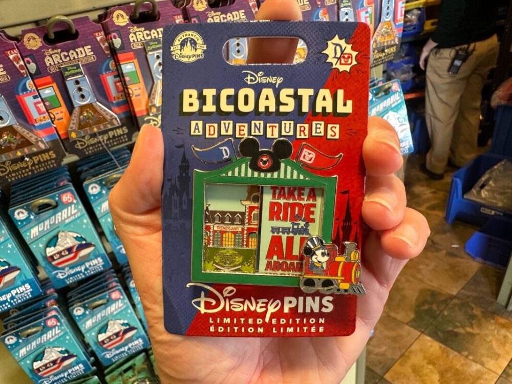 A hand holds a Disney Bicoastal Adventures limited edition Railroad pin featuring a "Take a Ride" design. The pin is displayed in its packaging with Disney branding.