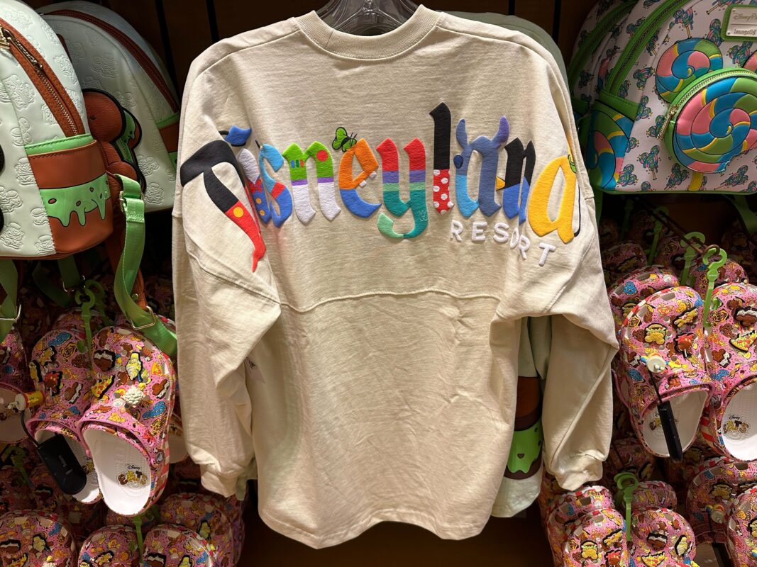 A white sweatshirt with "Disneyland Resort" written in colorful, playful letters is displayed on a hanger, surrounded by Disney-themed merchandise including backpacks and clogs.