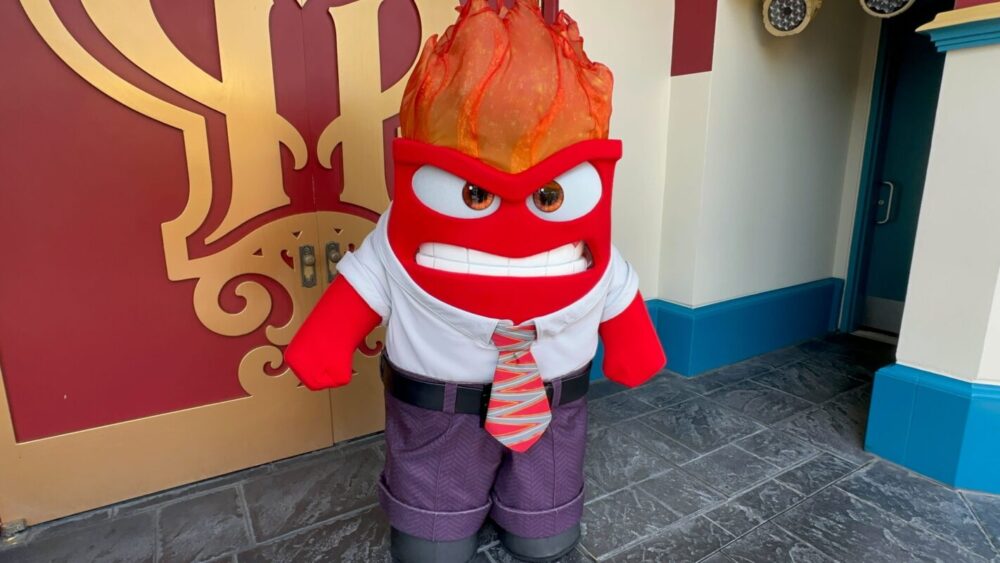 A person in a costume of the character "Anger" from the movie "Inside Out" stands in front of a decorative wall. The character has red skin, a fiery head, and is wearing a white shirt and tie.