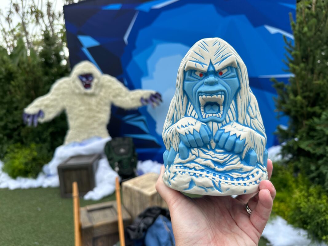 A blue Yeti figurine is held in the foreground with a larger Yeti decoration set against a blue backdrop in the background. Trees and snow-like decor surround the scene.
