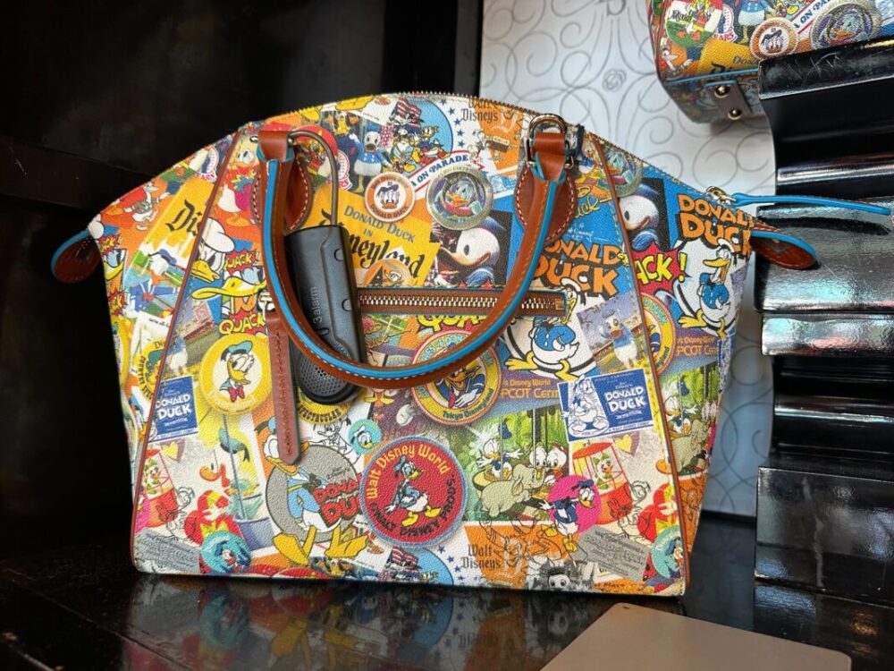 A handbag with various colorful Disney-themed prints, including characters and logos like Donald Duck and Disneyland. The bag has brown leather handles and a small visible pocket with a black item inside.