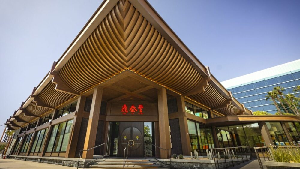 A modern building with prominent wooden architectural features, large glass windows, and red Chinese characters on the entrance. The building has a triangular roof and is surrounded by a clear sky.