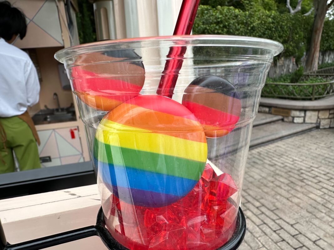 A clear plastic cup containing red plastic gems, rainbow-colored lollipops, and a drink with a red straw, placed on a counter outdoors. A person and a green garden background are partially visible.