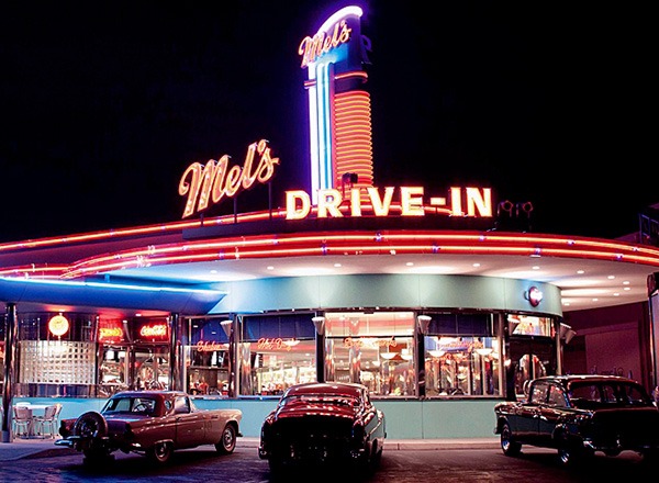 Night view of Mel's Drive-In featuring neon lights and vintage cars parked outside the retro-themed diner, reminiscent of a Universal Studios Picture.