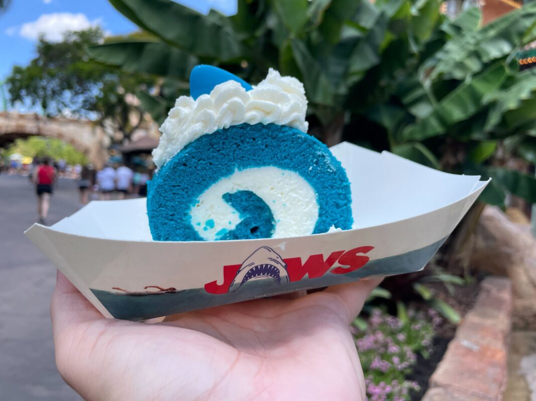 A hand holds a blue and white shark-themed cake roll in a "Jaws" branded container from Universal Orlando Resort. Trees and people can be seen in the background.