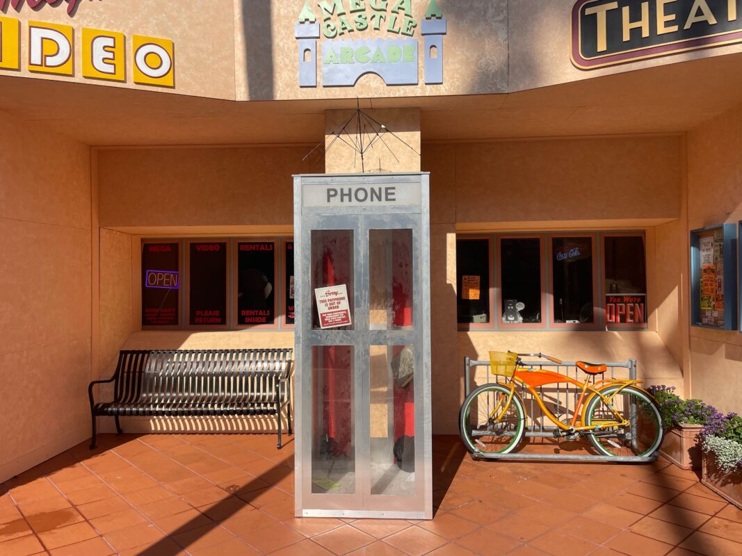 A glass phone booth stands in front of a retro-styled building with signs for an arcade, theater, and video store. To the right of the booth, a yellow bike is parked, and a bench is on the left.